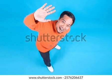 Full Length High Angle View Of Emotional Young Man Waving Hand To Camera On Blue Studio Wall 