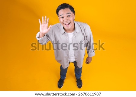 Full Length High Angle View Of Excited Young Man Waving Hand To Camera On Yellow Studio Wall