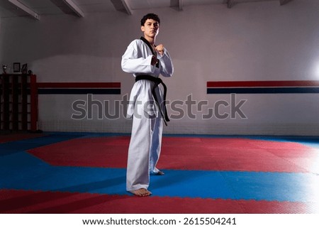 Full length of hispanic man practicing taekwondo wearing dobok and black belt, posing in fighting stance inside dojo