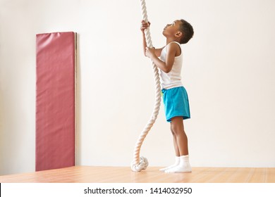 Full length image of handsome skinny African American schoolboy wearing white socks, t-shirt and blue shorts standing on mat during physical education class, looking up, going to climb rope