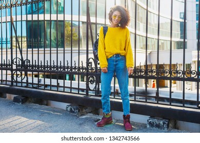 Full length Image of hipster African woman standing on  modern  buildings background. Wearing yellow cardigan and blue jeans. Spring fashion look.