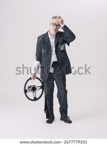 Full length image of a man in torn clothes holding a car steering wheel with one hand and touching his head with the other, looking sad and worried. Studio shot on white background.
