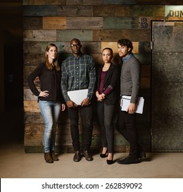 Full length image of multiracial creative team posing for camera in office. Young business people standing together against a wooden wall.