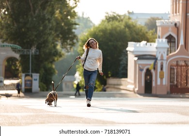 Vollständiges Bild einer hübschen, glücklichen Brunette-Frau in Herbstkleidung und Brille, die mit ihrem Hund im Park spaziert