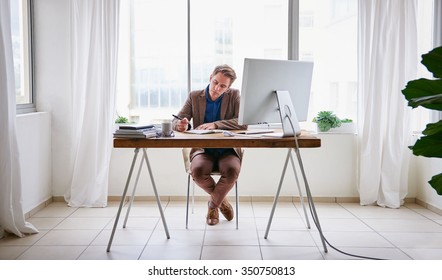 Full Length Image Of A Young Male Entrepreneur Sitting At His Desk In A Contemporary Work Space For One Person And Writing In His Journal