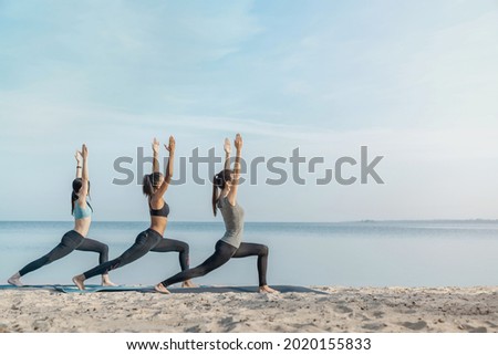 Full length image of young multiethnic group of woman practicing yoga exercise at the beach near water. Stretching exercises, active lifestyle, fitness, pilates, workout outdoors.