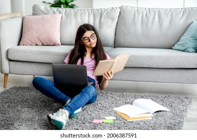 Full Length Of Indian Teen Girl Reading Textbook, Sitting On Floor With Laptop And Study Materials, Learning Remotely From Home, Copy Space. Focused Adolescent Getting Ready For Online Webinar