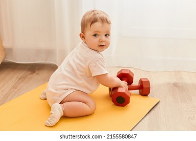 Full Length Indoor Shot Of Charming Cute Infant Baby Crawling On Yoga Mat With Dumbbells, Exploring Sport Equipment, Wearing Bodysuit.