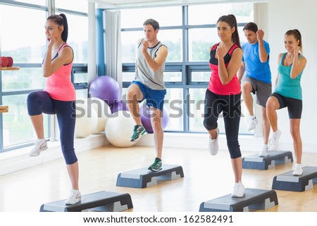 Full length of instructor with fitness class performing step aerobics exercise in gym