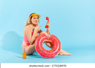 Full length, irritated resentful woman in bathing suit sitting with rubber ring and pointing at phone handset, grimacing madly, looking at camera with vexed annoyed expression. studio shot isolated