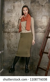 Full length isolated studio portrait of a young Asian lady in an olive skirt, a beige jacket and a terra cotta scarf. The brunette girl posing against the grunge rusty background, looking to the side.