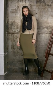 Full length isolated studio portrait of a young Asian lady in an olive skirt, a beige jacket and a black scarf. The brunette girl posing against the grunge rusty background, looking at the camera.