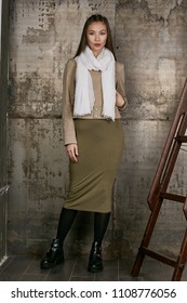 Full length isolated studio portrait of a young Asian lady in an olive skirt, a beige jacket and a white scarf. The brunette girl posing against the grunge rusty background, looking at the camera.