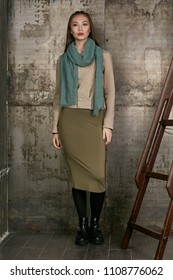 Full length isolated studio portrait of a young Asian lady in an olive skirt, a beige jacket and a sage scarf. The brunette girl posing against the grunge rusty background, looking at the camera.