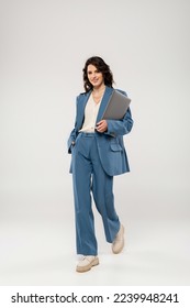 full length of joyful woman in fashionable suit holding hand in pocket while walking with laptop on grey background - Shutterstock ID 2239948241