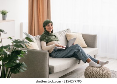 Full length of joyful young Arab woman in hijab working on laptop, sitting on couch at home office, copy space. Cheery Middle Eastern lady having business webinar or remote meeting on pc - Powered by Shutterstock