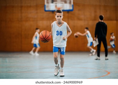 Full length of a junior basketball player in action dribbling a ball towards camera at indoor court on his training. In blurry background is his team practicing basketball.Athlete dribbling basketball - Powered by Shutterstock