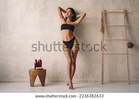Full length of korean woman in black bikini posing near textured wall in the bathroom
