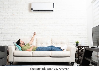 Full Length Of Latin Man Turning On Mini Split While Relaxing On Sofa At Home