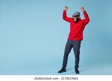 Full length laughing joyful young bearded african american man in casual red shirt cap eyeglasses standing doing winner gesture clenching fists isolated on pastel blue wall background studio portrait