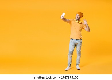 Full Length Of Laughing Young Bearded Man Wearing Basic Casual T-shirt Headphones Hat Doing Selfie Shot On Mobile Phone Waving Greeting With Hand Isolated On Bright Yellow Background, Studio Portrait