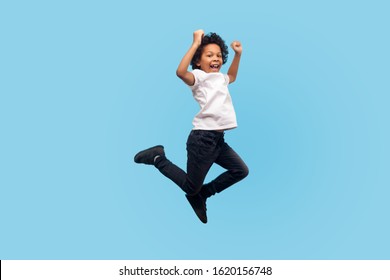 Full length, lively energetic little boy in T-shirt and denim jumping in air screaming with happiness, child flying up, feeling inspired crazy and overjoyed. indoor studio shot, blue background