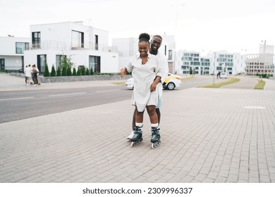 Full length of loving African American boyfriend in casual clothes teaching smiling girlfriend in rollerblades skate during date on paved city street