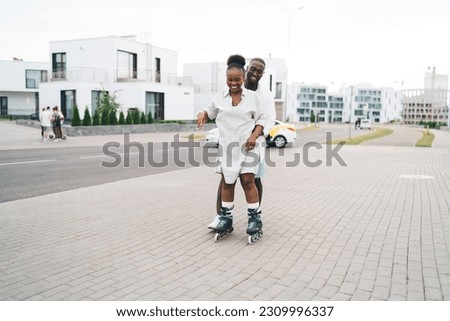 Full length of loving African American boyfriend in casual clothes teaching smiling girlfriend in rollerblades skate during date on paved city street