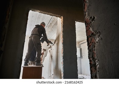 Full Length Of Male Builders Using Hammer Drill And Sledgehammer While Destroying Wall In Building. View Through Doorway Of Two Workers Breaking Wall In Apartment Under Renovation.