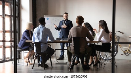 Full Length Male Speaker Presenting Project Financial Analysis Or Corporate Marketing Strategy On Flipchart To Focused Managers At Office. Presenter Holding Educational Workshop To Diverse Colleagues.