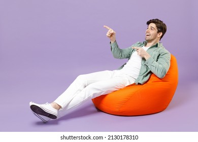 Full length man 20s wear casual mint shirt white t-shirt sitting in orange bean bag chair point index finger aside on workspace area isolated on purple color background studio People lifestyle concept - Shutterstock ID 2130178310