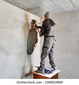 Full Length Of Man Builder Standing On Wooden Table And Breaking Wall With Hammer While Working On Home Refurbishment. Male Worker Holding Sledgehammer And Destroying Wall In Room Under Renovation.