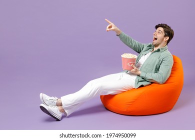 Full Length Man In Casual Mint Shirt White T-shirt Sitting In Orange Bean Bag Chair Hold Takeaway Bucket Eat Popcorn Watch Movie Film Point Finger Aside Isolated On Purple Background Studio Portrait.