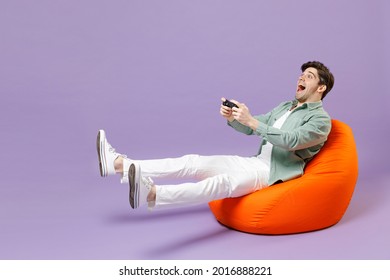 Full Length Man In Casual Mint Shirt White T-shirt Sitting In Orange Bean Bag Chair Hold Takeaway Bucket Eat Popcorn Watch Movie Film Play Pc Game With Joystick Console Isolated On Purple Background.