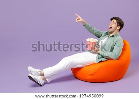 Full length man in casual mint shirt white t-shirt sitting in orange bean bag chair hold takeaway bucket eat popcorn watch movie film point finger aside isolated on purple background studio portrait.