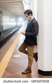 full length of man in medical mask standing and holding smartphone in hall of subway