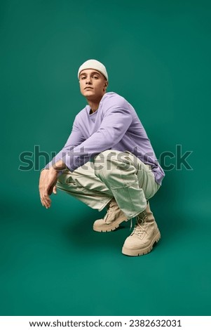 full length of man in purple sweatshirt and beanie hat sitting on turquoise backdrop, winter style