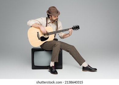 full length of man retro style clothing playing acoustic guitar and sitting on antique tv on grey