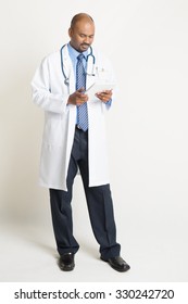 Full Length Mature Indian Male Medical Doctor In Uniform Using Digital Tablet Pc, Standing On Plain Background With Shadow.