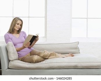 Full Length Of A Middle Aged Woman Reading Book On Sofa At Home