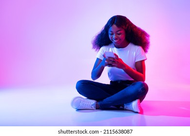 Full Length Of Millennial African American Lady Sitting Cross Legged With Cellphone, Surfing Social Media, Chatting To Friend In Neon Light. Cool Black Woman Using Mobile Device, Free Space