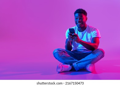 Full Length Of Millennial African American Guy Sitting Cross Legged With Smartphone, Surfing Social Media, Chatting To Friend In Neon Light, Copy Space. Cool Black Man Using Mobile Phone