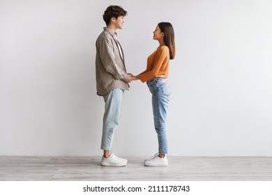 Full length of millennial Asian couple holding hands and looking at each other against white studio wall, side view. Loving young male and female demonstrating their love