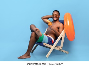 Full Length Of Millennial Black Guy Drinking Tropical Cocktail In Lounge Chair Over Blue Studio Background. Happy African American Man Chilling On Beach With Refreshing Drink During Summer Vacation