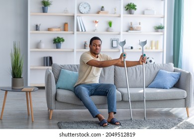 Full Length Of Millennial Black Guy Having Difficulty Standing Up From Couch, Using Crutches At Home. 