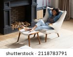 Full length mindful carefree young African American woman napping in comfortable armchair with legs on ottoman, breathing fresh air, sleeping in modern living room, enjoying peaceful weekend time.
