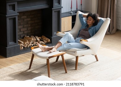 Full length mindful carefree young African American woman napping in comfortable armchair with legs on ottoman, breathing fresh air, sleeping in modern living room, enjoying peaceful weekend time.