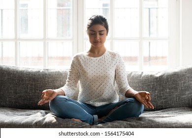 Full length mindful young indian woman making mudra gesture, sitting in lotus position on comfortable couch at home. Peaceful millennial girl deeply meditating, doing breathing yoga exercises alone.