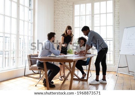 Full length motivated young diverse teammates gathering near table in modern office, working on corporate growth strategy development, analyzing paper marketing research report, discussing ideas.