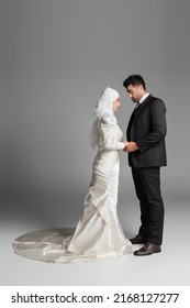 full length of muslim bride in wedding dress and groom in suit holding hands on grey